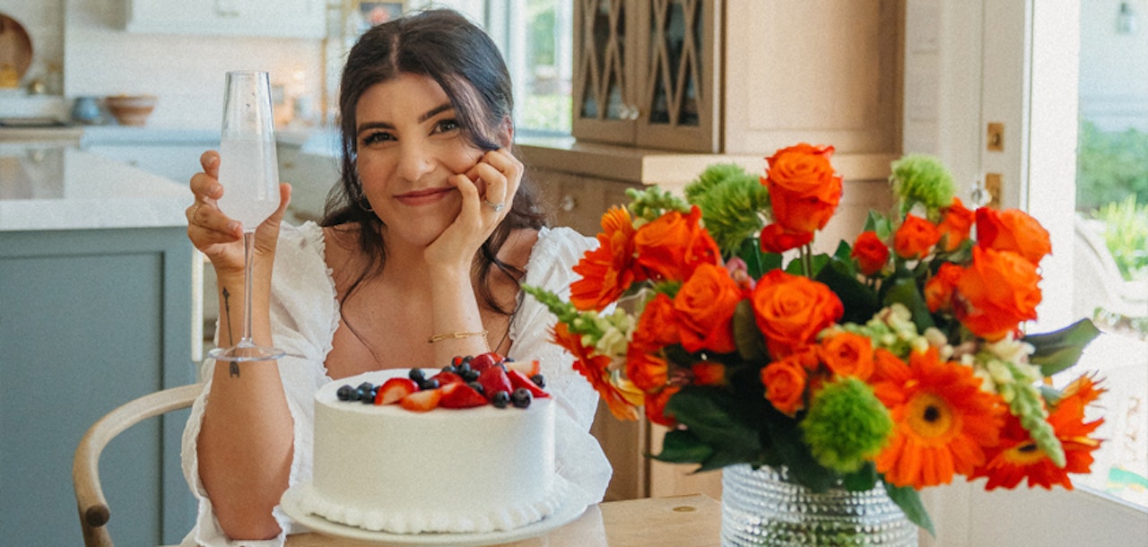 A cheerful woman toasts with a drink, celebrating beside a vibrant cake and a colorful floral arrangement.