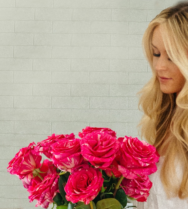 A woman with long blonde hair holding a vibrant bouquet of pink roses against a textured wall.