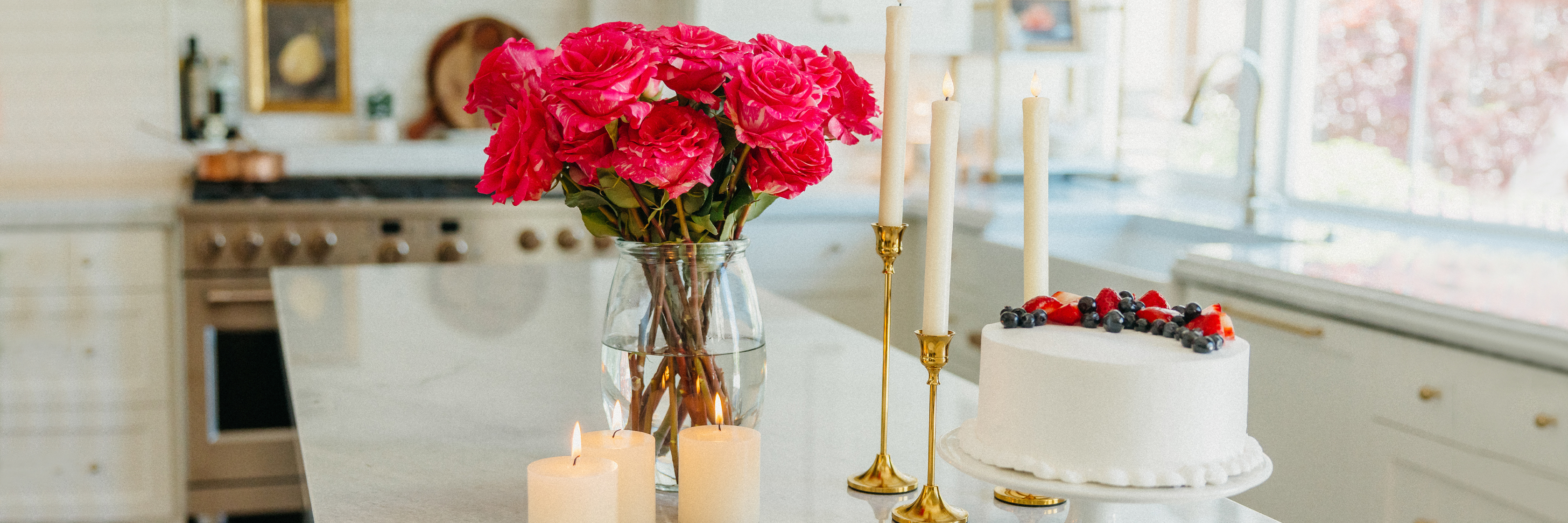Bright pink roses in a vase on a kitchen island, accompanied by candles and a decorated cake.