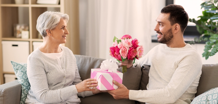 A smiling young man giving a pink gift box and a floral bouquet to an older woman at home.