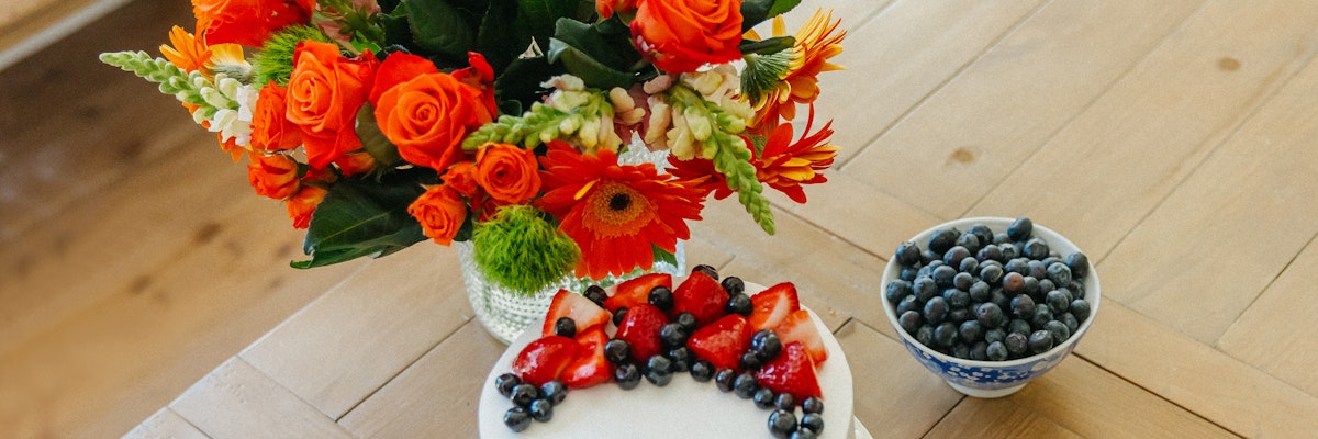 Vibrant floral arrangement with orange roses and colorful blooms beside a fresh berry-topped cake.