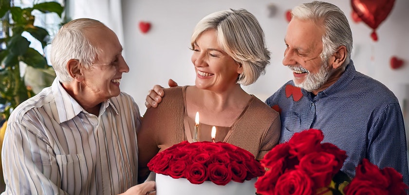A joyful celebration with a woman surrounded by two smiling men, holding a beautiful rose cake.