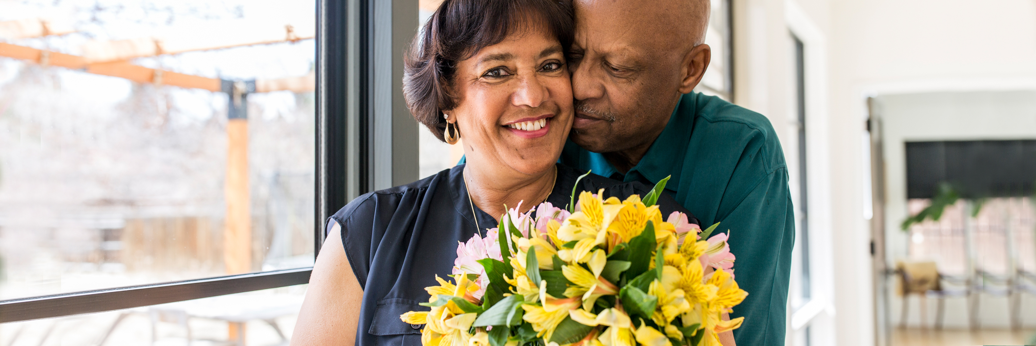 Couple sharing a joyful moment while holding a vibrant yellow and pink floral bouquet indoors.