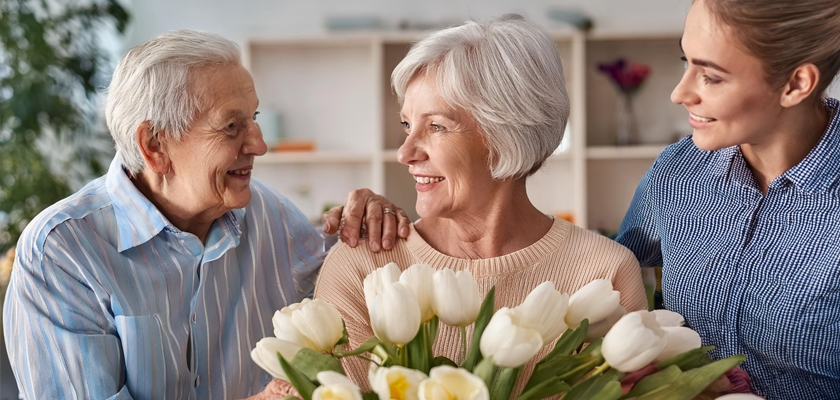 A heartwarming scene of two women smiling at an elderly woman holding a bouquet of white tulips.