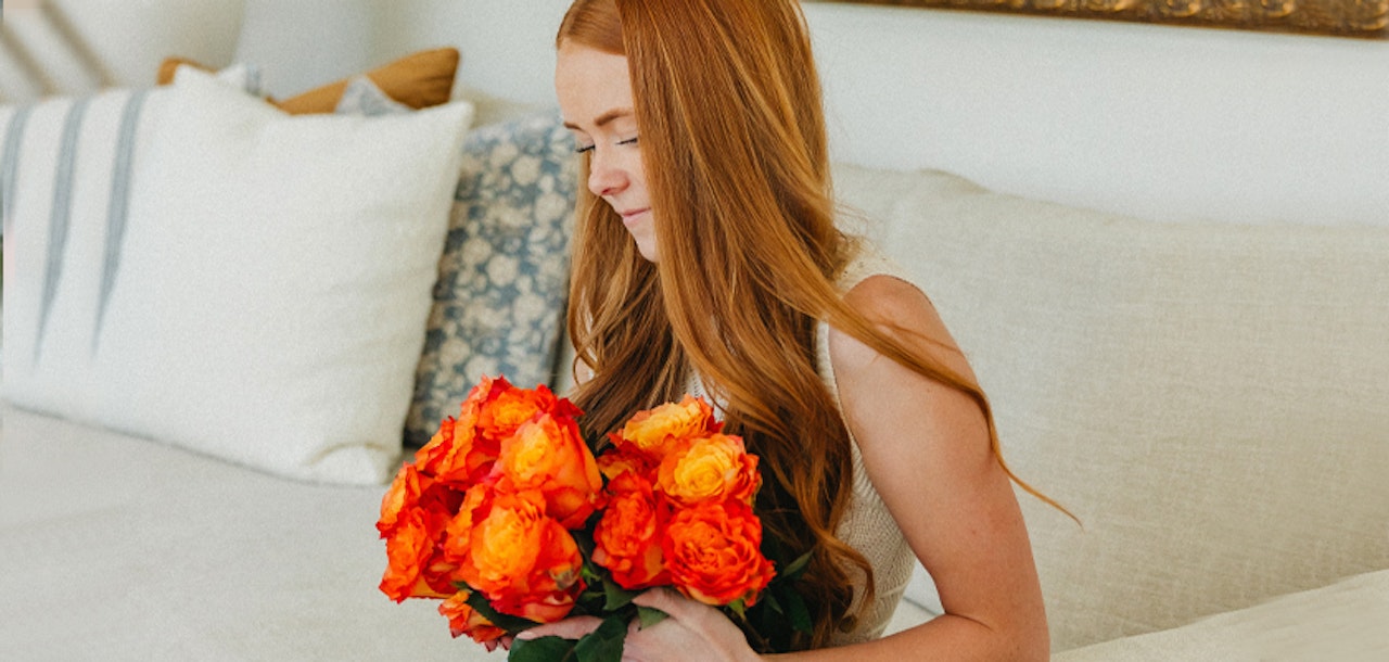 A woman with long red hair holding a vibrant bouquet of orange roses, seated on a cozy sofa.