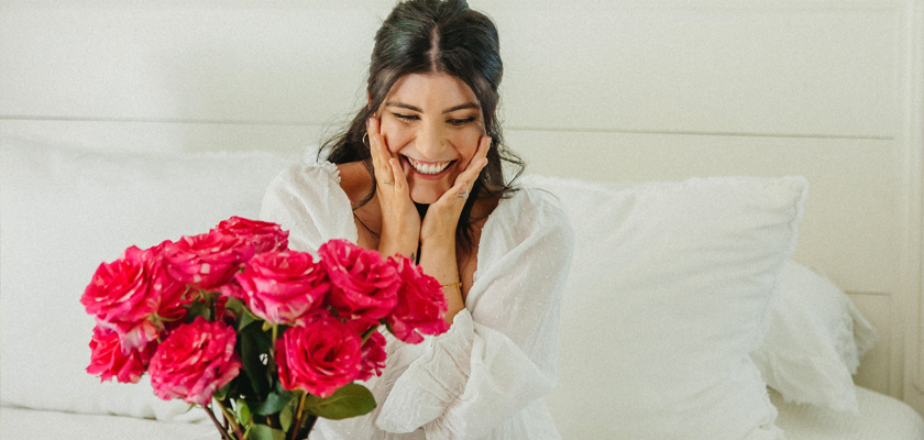 A joyful woman admiring a vibrant bouquet of pink roses in a bright, elegant setting.