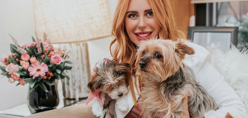 Woman with long blonde hair smiling while holding two small dogs, surrounded by flowers.