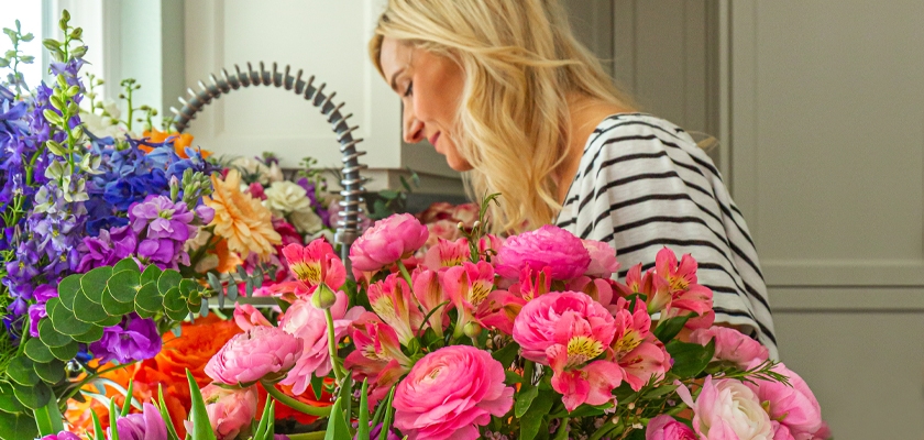A woman arranging a colorful bouquet of pink and purple flowers in a bright and cheerful workspace.