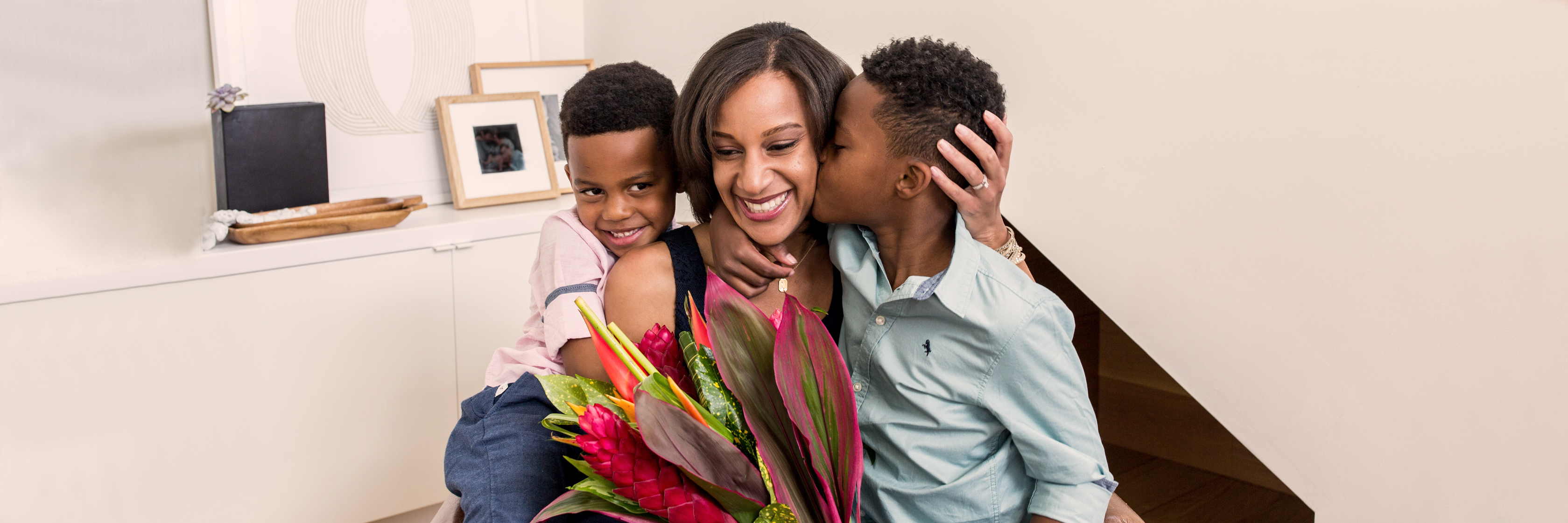 A joyful woman embraces her two children, surrounded by vibrant flowers in a cozy home setting.