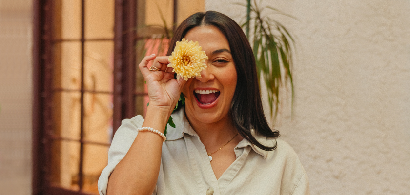 A joyful woman holds a yellow flower to her eye, smiling brightly in a cozy indoor setting.
