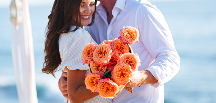 A couple embracing on the beach, holding a vibrant bouquet of peach roses, symbolizing love and joy.
