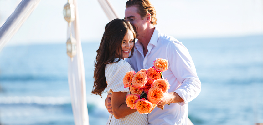 A joyful couple on the beach, sharing a moment with a bouquet of peach-colored roses.