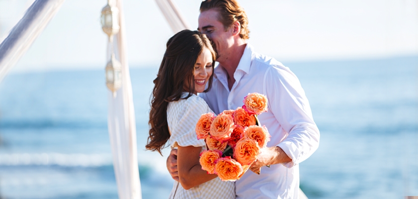A joyful couple on the beach, sharing a moment with a bouquet of peach-colored roses.