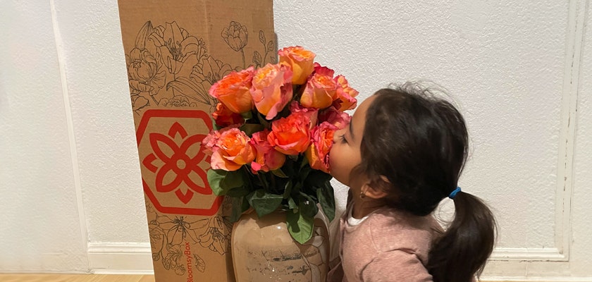 Little girl playfully smelling a vibrant bouquet of orange roses beside a floral delivery box.