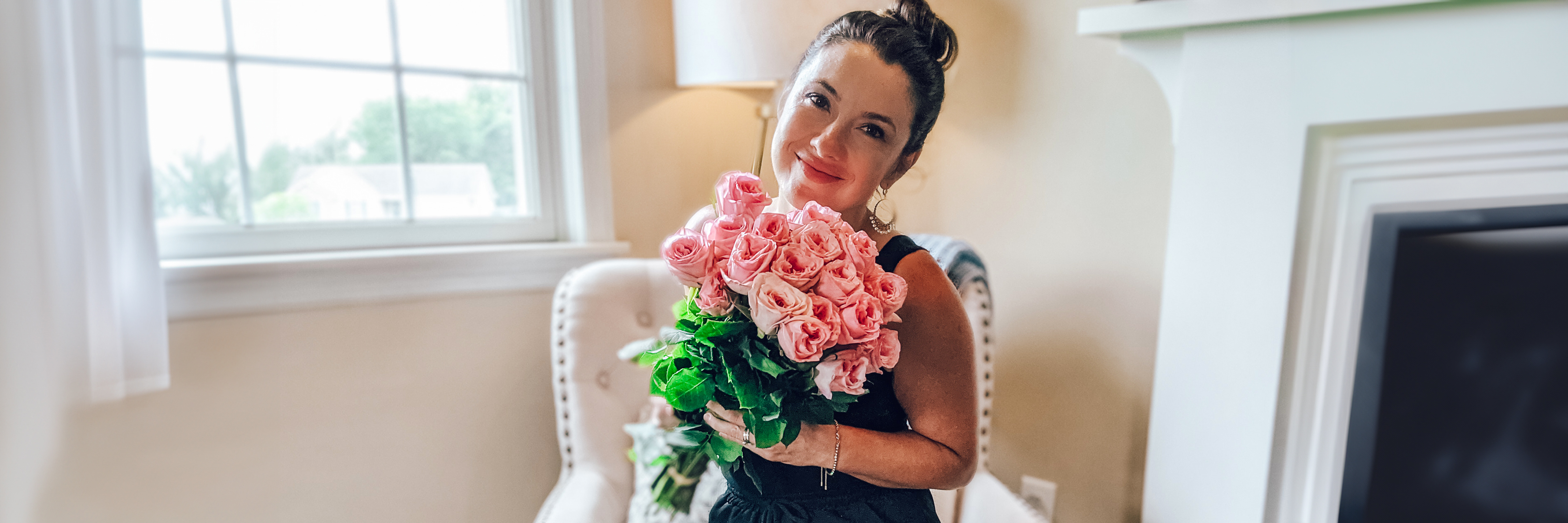 A smiling woman holds a lush bouquet of pink roses in a cozy room, radiating warmth and joy.