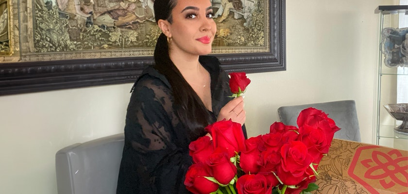A woman holding a red rose, seated beside a stunning bouquet of roses, radiating elegance and charm.