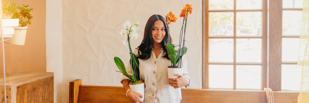 A woman holding two vibrant potted orchid plants, smiling in a bright indoor setting.