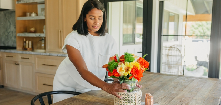A woman arranging a vibrant floral centerpiece on a rustic wooden table in a bright kitchen setting.
