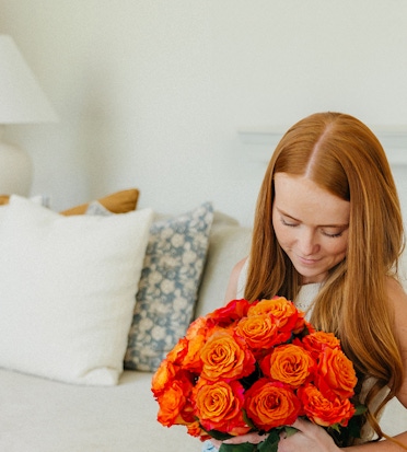 A woman holds a vibrant bouquet of orange roses, seated comfortably on a cozy couch.