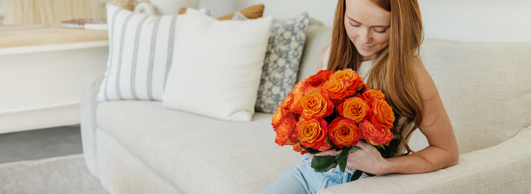 A woman admiring a vibrant bouquet of orange roses while seated on a cozy sofa.