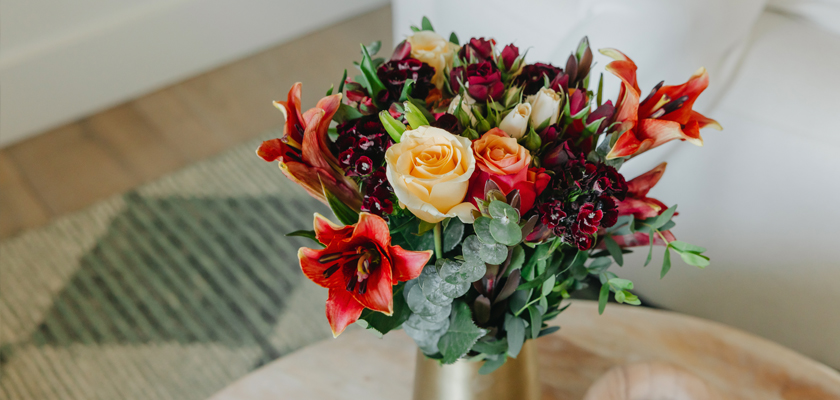 Vibrant bouquet of roses, lilies, and greenery arranged elegantly in a gold vase.