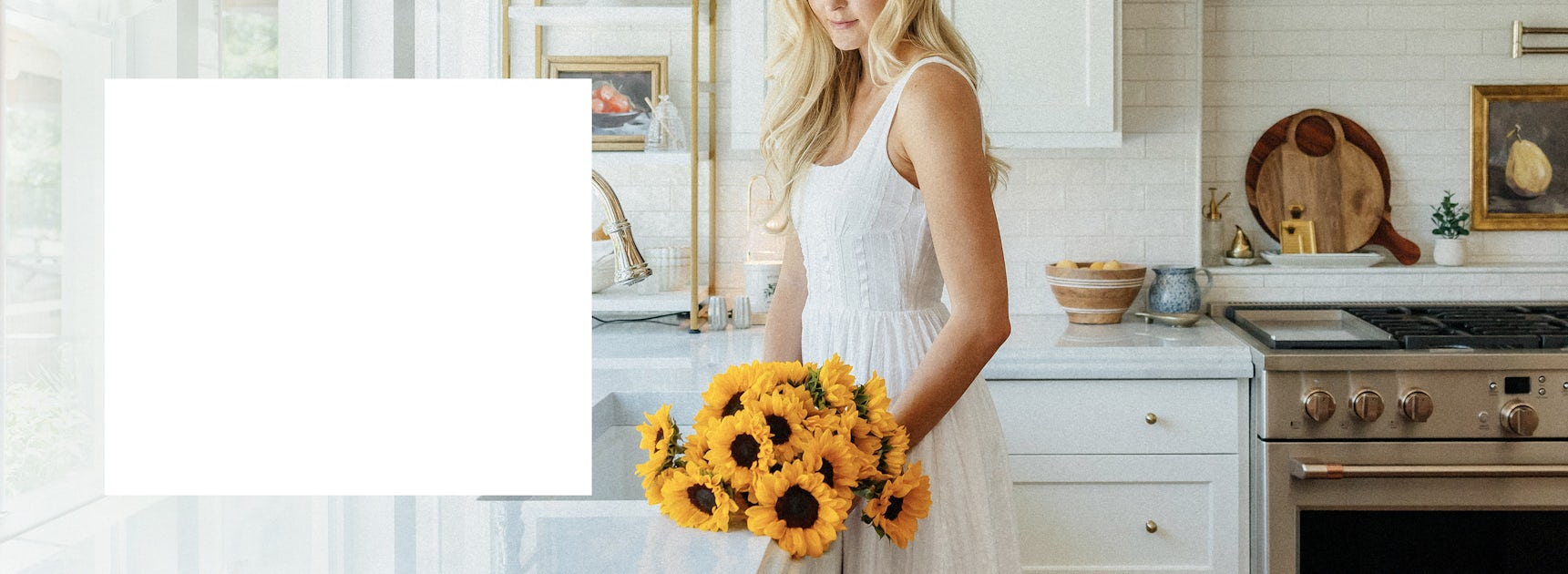 Woman in a white dress holding a vibrant bouquet of sunflowers in a bright, airy kitchen.