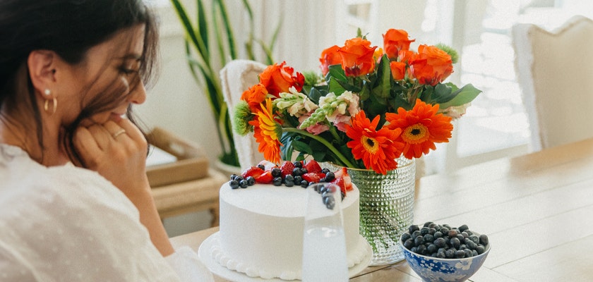 A woman enjoying a celebration with a white cake, vibrant flowers, and fresh berries on the table.