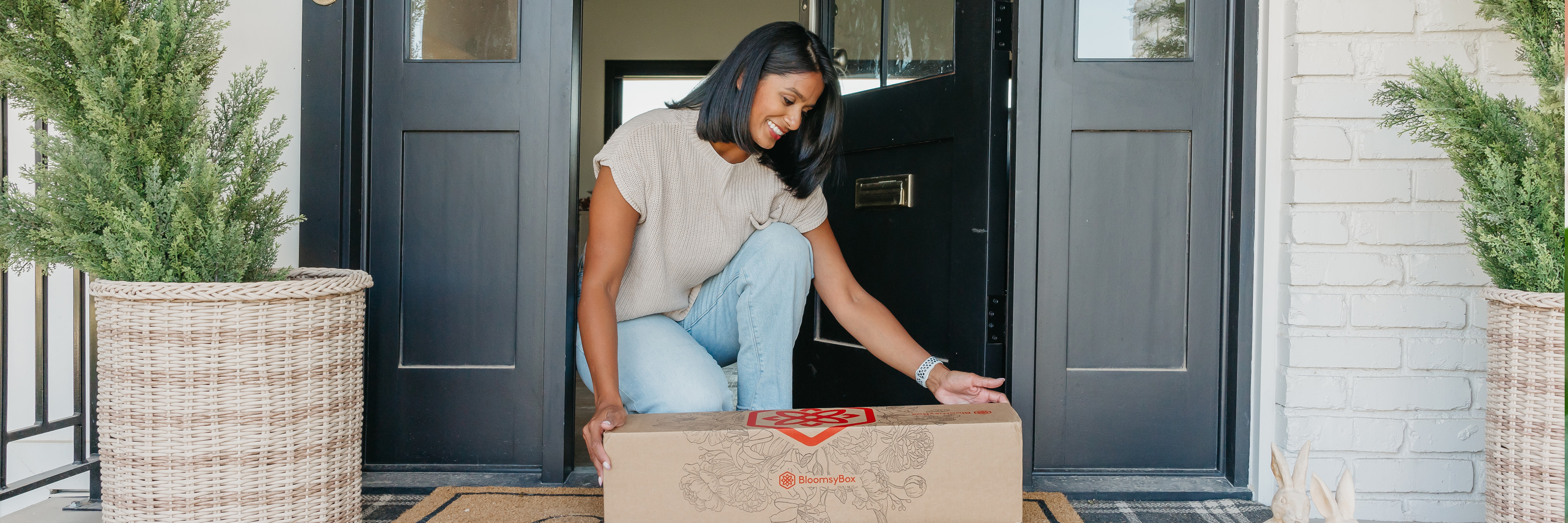 A woman joyfully receives a floral delivery package on her doorstep, surrounded by greenery.
