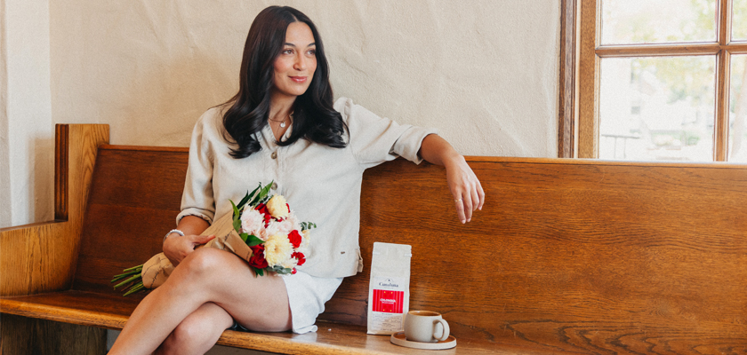 A woman sits on a wooden bench, holding a vibrant bouquet of flowers beside a coffee cup and bag.