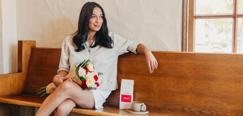 A woman sits on a wooden bench, holding a vibrant bouquet of flowers beside a coffee cup and bag.