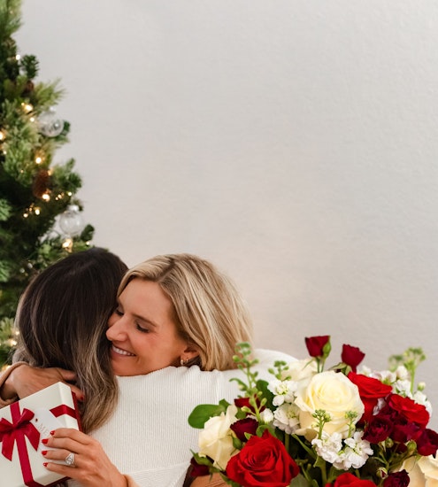 Two women joyfully embracing, surrounded by a festive floral bouquet of red and white roses.
