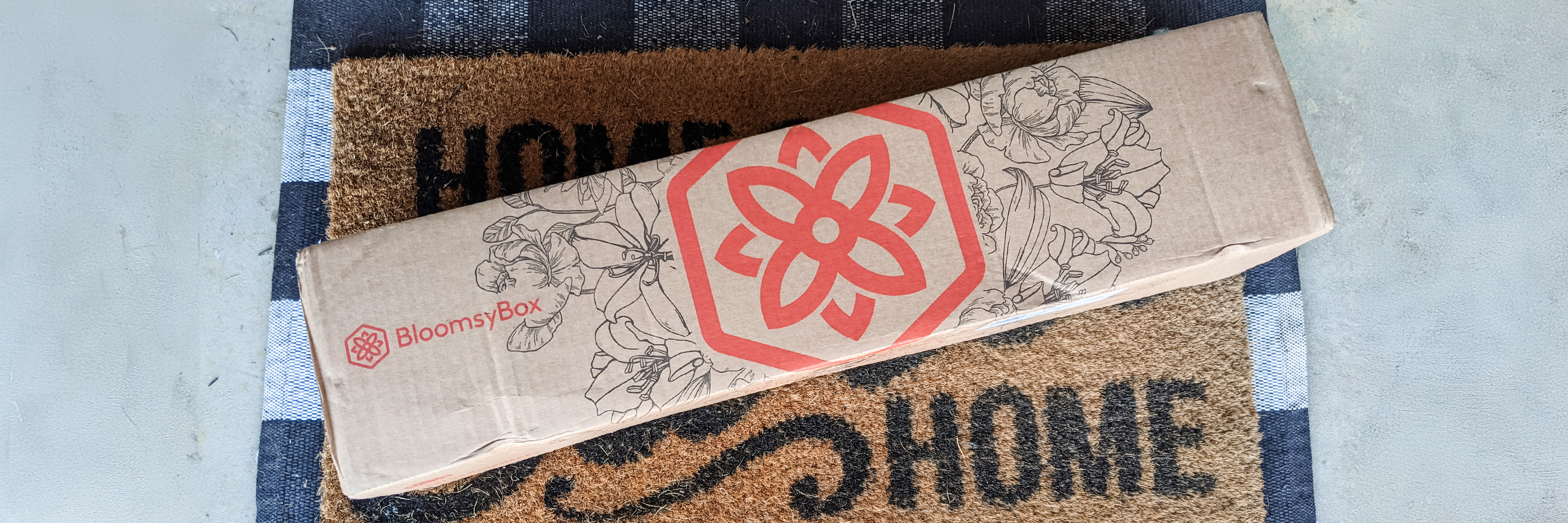 A BloomsyBox package featuring a floral design, resting on a welcome mat at a home entrance.