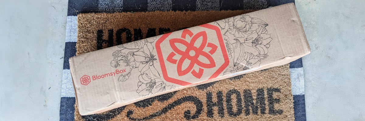 A BloomsyBox package featuring a floral design, resting on a welcome mat at a home entrance.