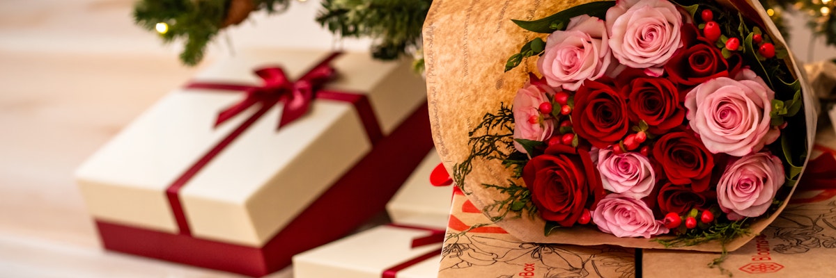 Festive bouquet of red and pink roses wrapped in paper, surrounded by holiday gifts.