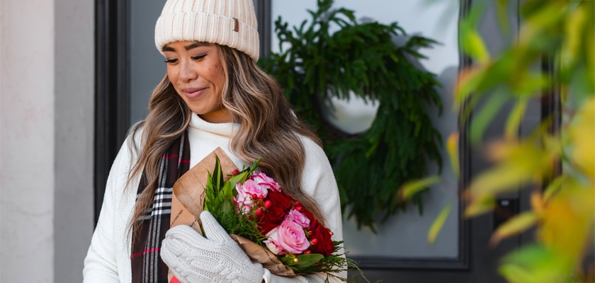 A woman in cozy winter attire holds a floral bouquet at her doorstep, surrounded by greenery.