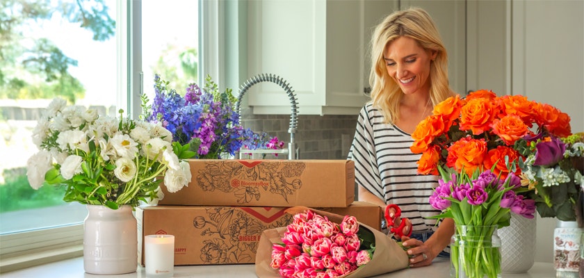 A woman arranging vibrant flowers in a bright kitchen, showcasing colorful blooms and gift boxes.