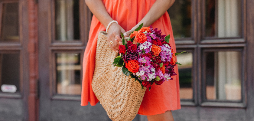 A joyful woman in an orange dress carries a vibrant floral bouquet in a woven basket.