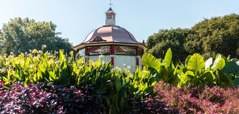 Charming gazebo surrounded by lush greenery and vibrant flowering plants on a sunny day.