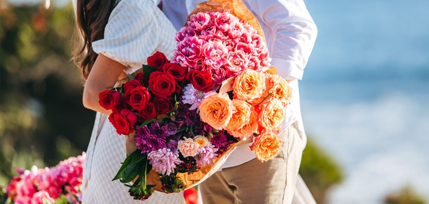 A couple embraces, holding a vibrant bouquet of roses and blooms by the ocean, capturing the moment.