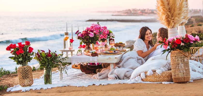 A romantic beach picnic setup with roses, vibrant flowers, and cozy blankets, perfect for couples.