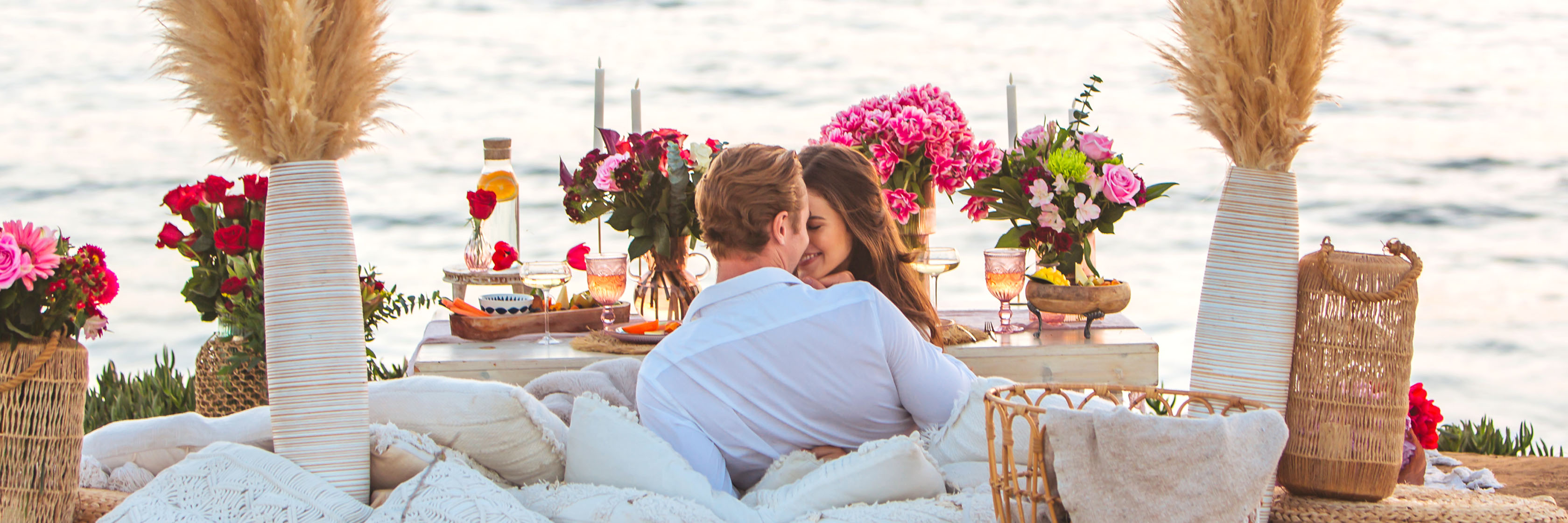 Couple enjoying a romantic picnic by the ocean, surrounded by beautiful floral arrangements.