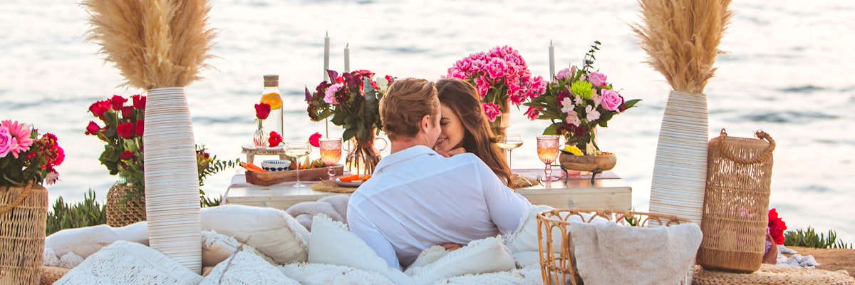 Couple enjoying a romantic picnic by the ocean, surrounded by beautiful floral arrangements.