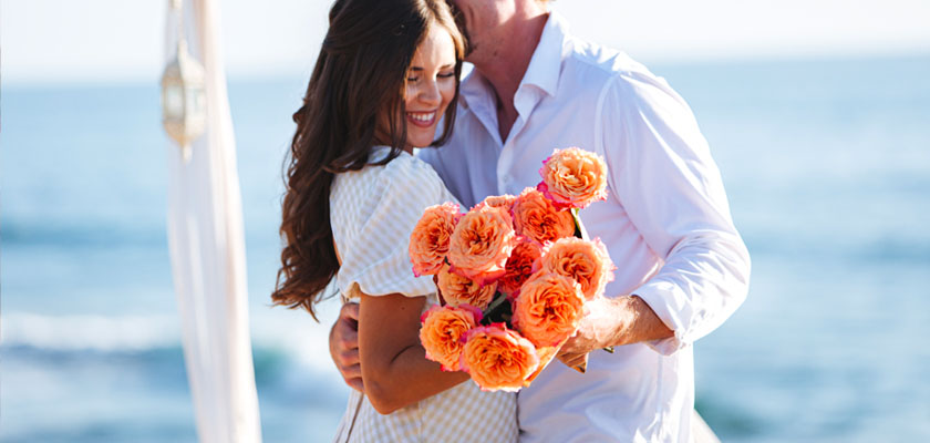 A joyful couple embracing on the beach, holding a vibrant bouquet of orange roses.