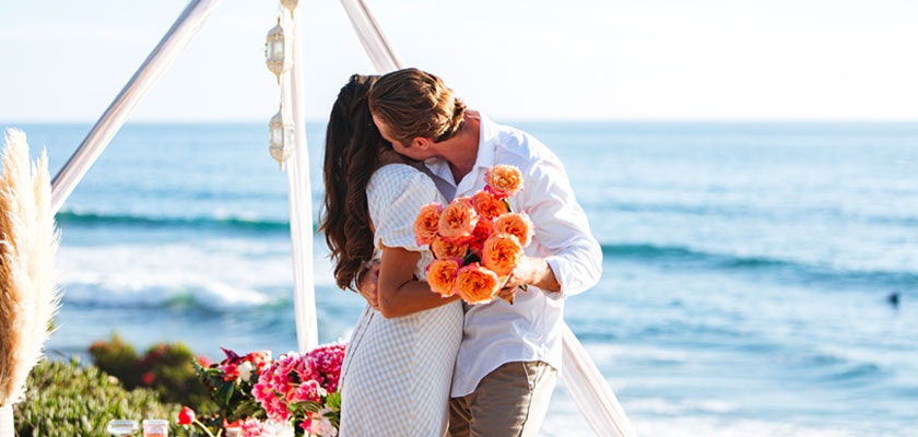A couple kisses passionately on the beach, surrounded by vibrant flowers in a romantic setting.