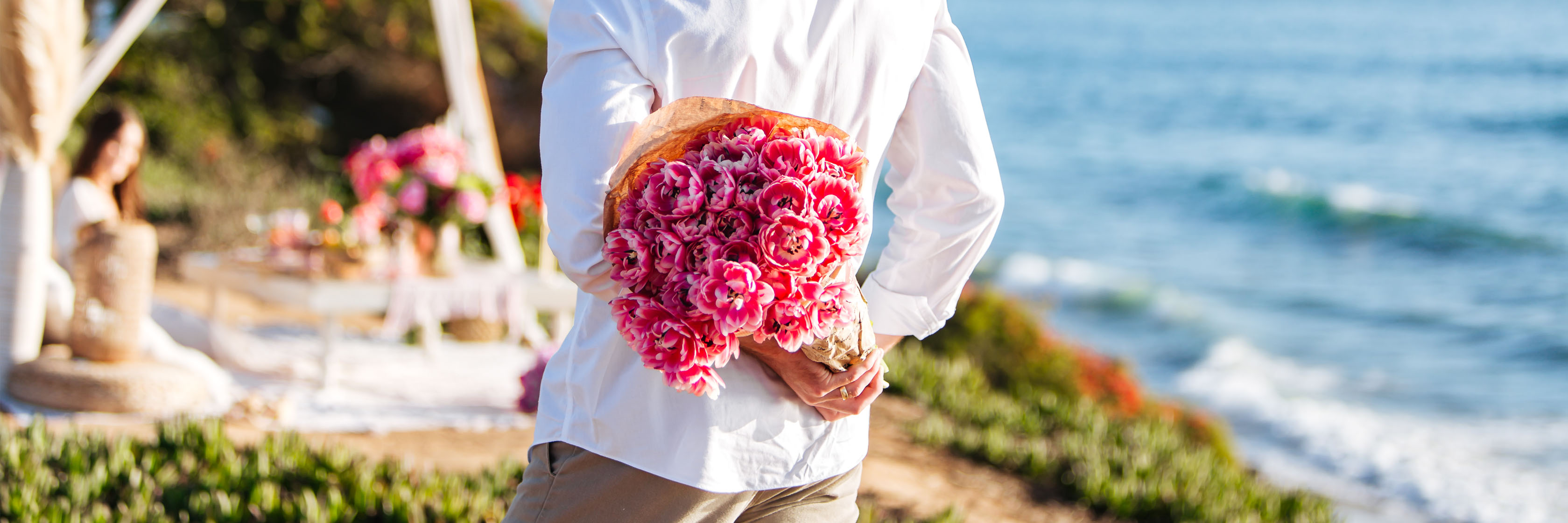A man holding a vibrant bouquet of pink flowers by the beach, perfect for romantic occasions.