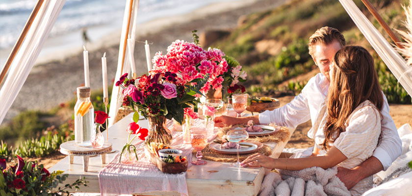 Couple enjoying a romantic beach picnic with flowers, candles, and gourmet food.