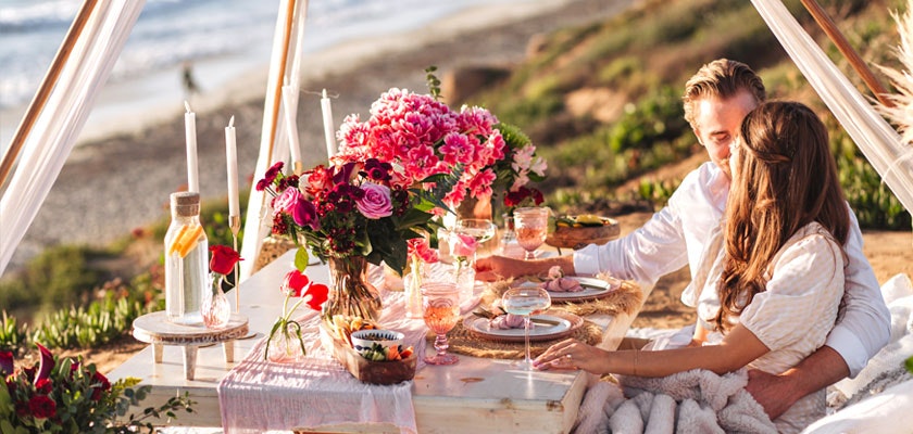 Couple enjoying a romantic beach picnic with flowers, candles, and gourmet food.