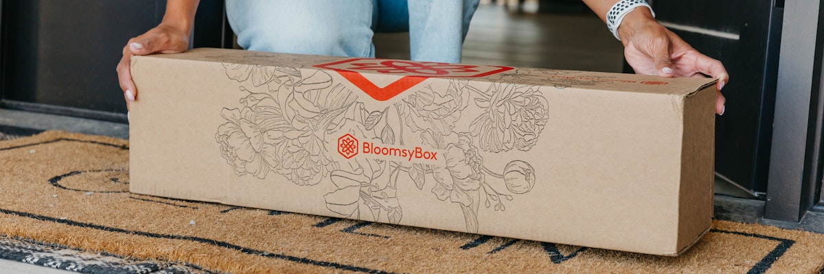 A person receiving a BloomsyBox floral delivery at their doorstep, showcasing beautiful blooms.