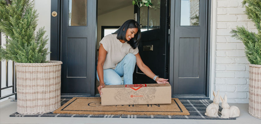 A woman smiling as she receives a floral subscription box at her door, surrounded by greenery.