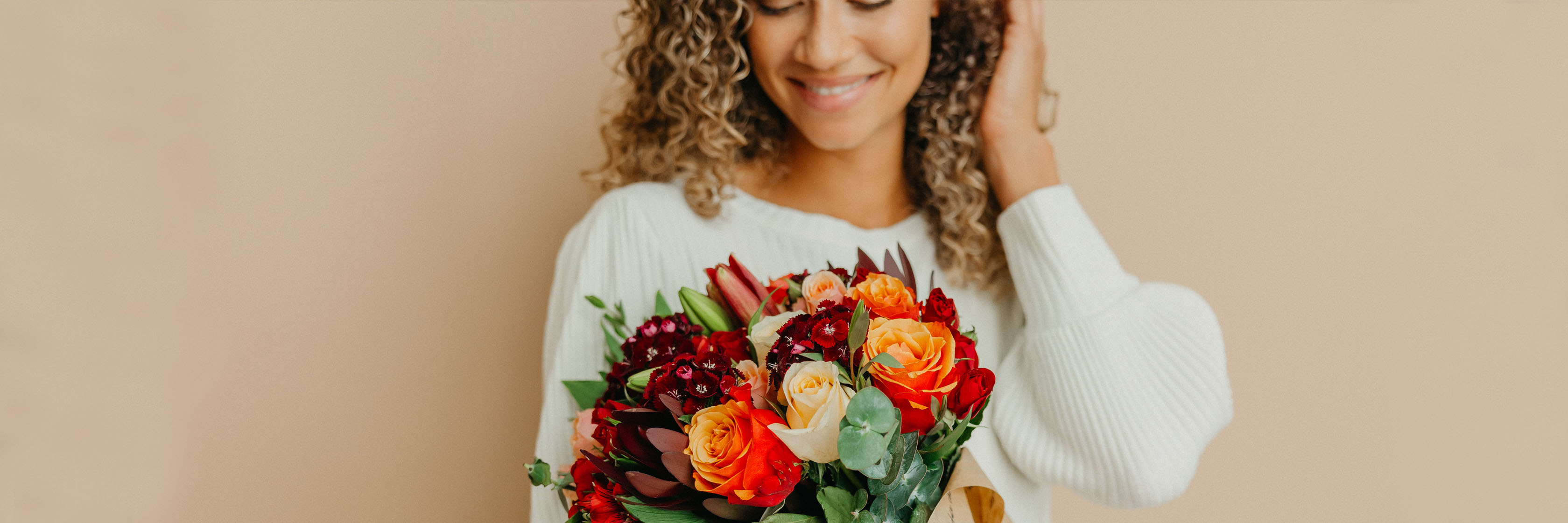 A smiling woman holds a vibrant bouquet featuring orange roses, red blooms, and lush greenery.